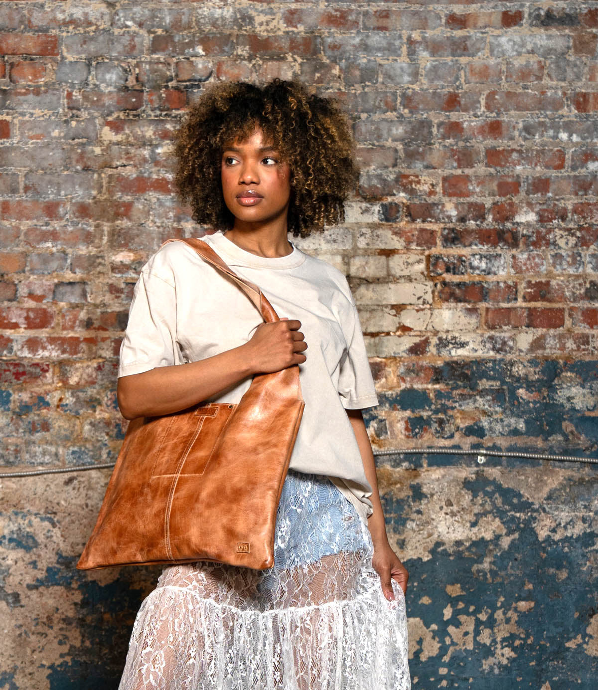 A person stands against a textured brick wall, wearing a beige t-shirt, sheer lace skirt, and carrying the Bed Stu Ariel II tote featuring a lightweight design and spacious compartment.