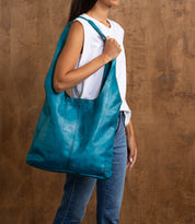 A person wearing a sleeveless white top and jeans stands against a brown background, holding the Ariel Max oversized tote in vibrant teal by Bed Stu.