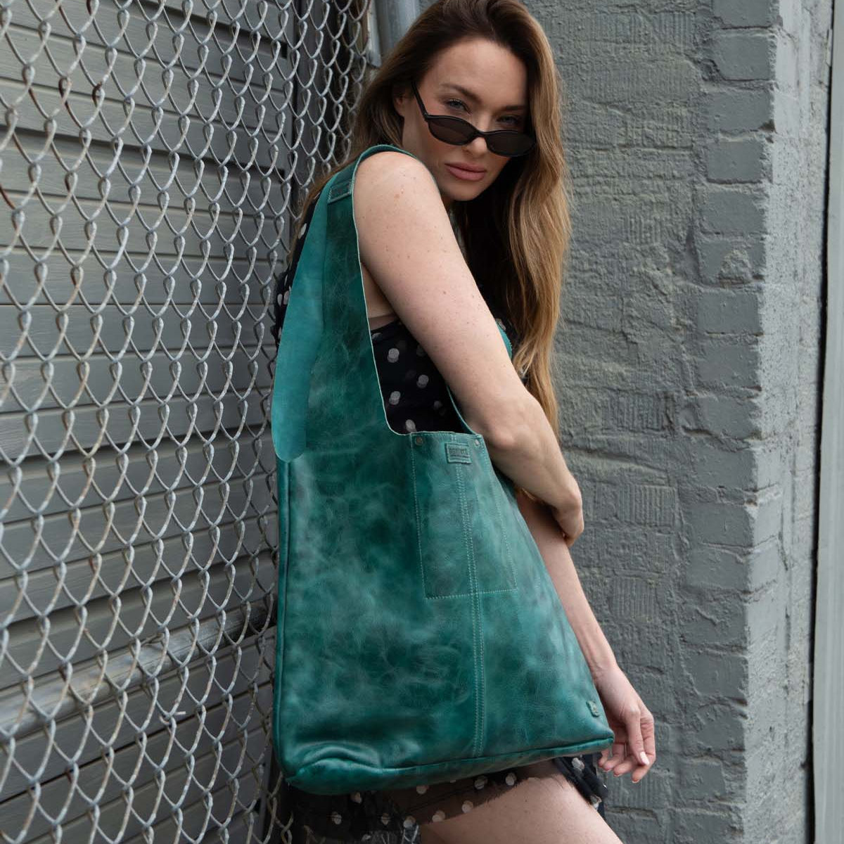A woman in a black dress with white spots poses by a chain-link fence, wearing sunglasses and carrying the Ariel Max by Bed Stu—an oversized artisanal tote bag in striking teal leather.