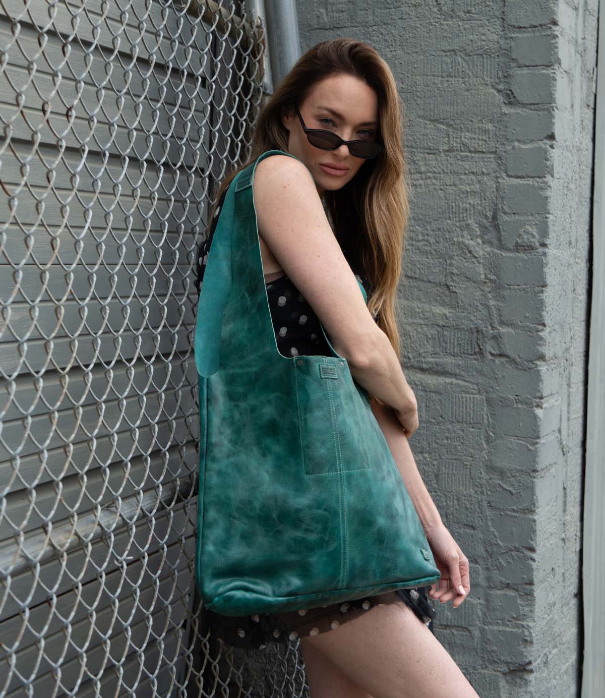 A woman in a black dress with white spots poses by a chain-link fence, wearing sunglasses and carrying the Ariel Max by Bed Stu—an oversized artisanal tote bag in striking teal leather.