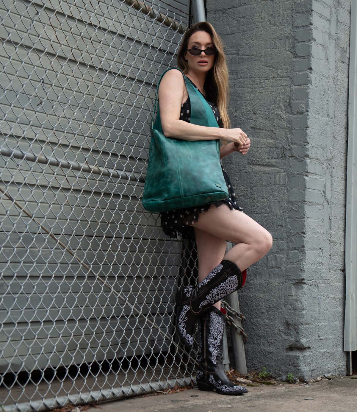 A woman in a short black dress with a white pattern, knee-high black cowboy boots, sunglasses, and the Bed Stu Ariel Max hand-finished leather oversized tote stands by a chain-link fence and gray wall.
