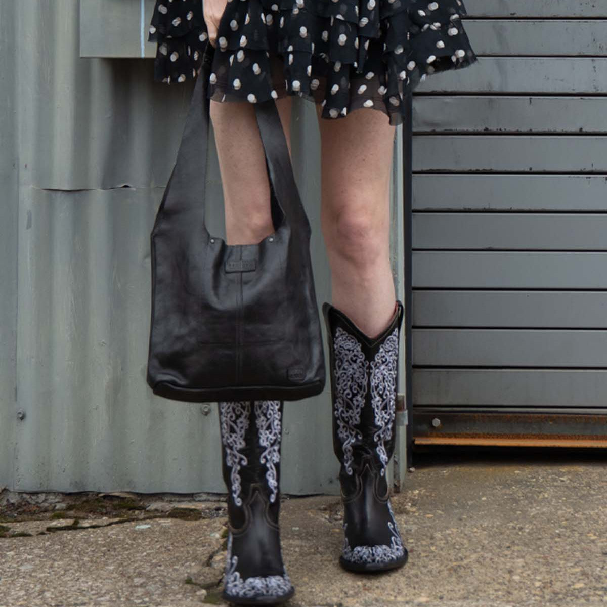 A person wearing a black and white skirt and embroidered cowboy boots holds the Bed Stu Ariel Mid hand-finished leather handbag, standing on concrete near a metal wall and garage door.
