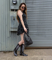 A woman in a sleeveless black polka dot dress, patterned cowboy boots, and sunglasses stands by a metal wall, holding the Bed Stu Ariel Mid— a hand-finished leather handbag with a magnetic closure.