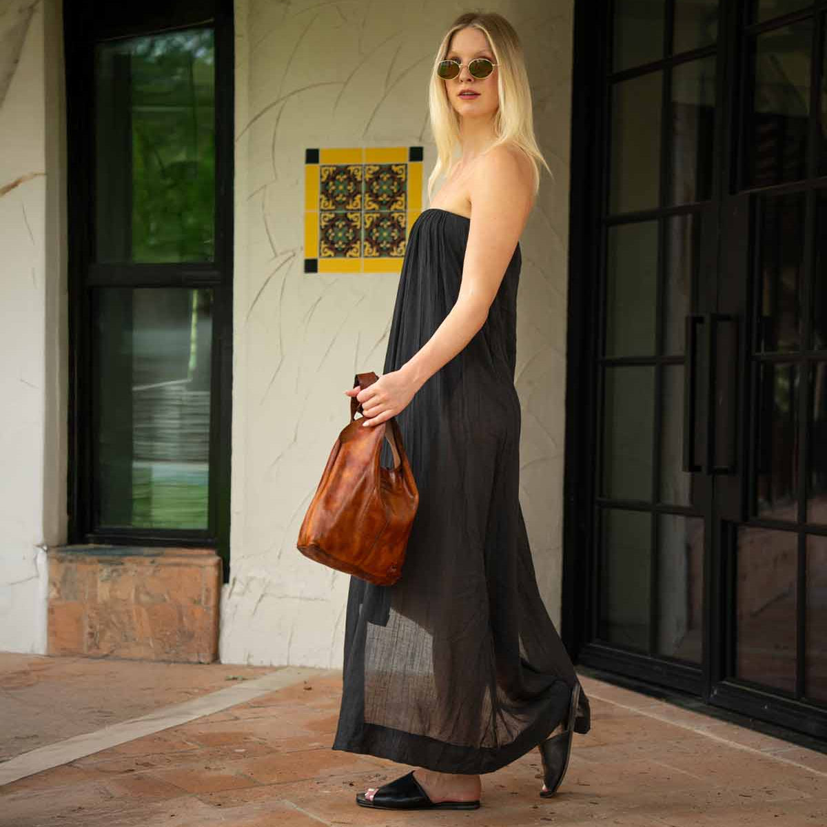 A woman in a black strapless dress, sunglasses, and sandals stands outdoors near glass doors, holding the Bed Stu Ariel Mid—an artisanal handbag crafted from vegetable-tanned leather.