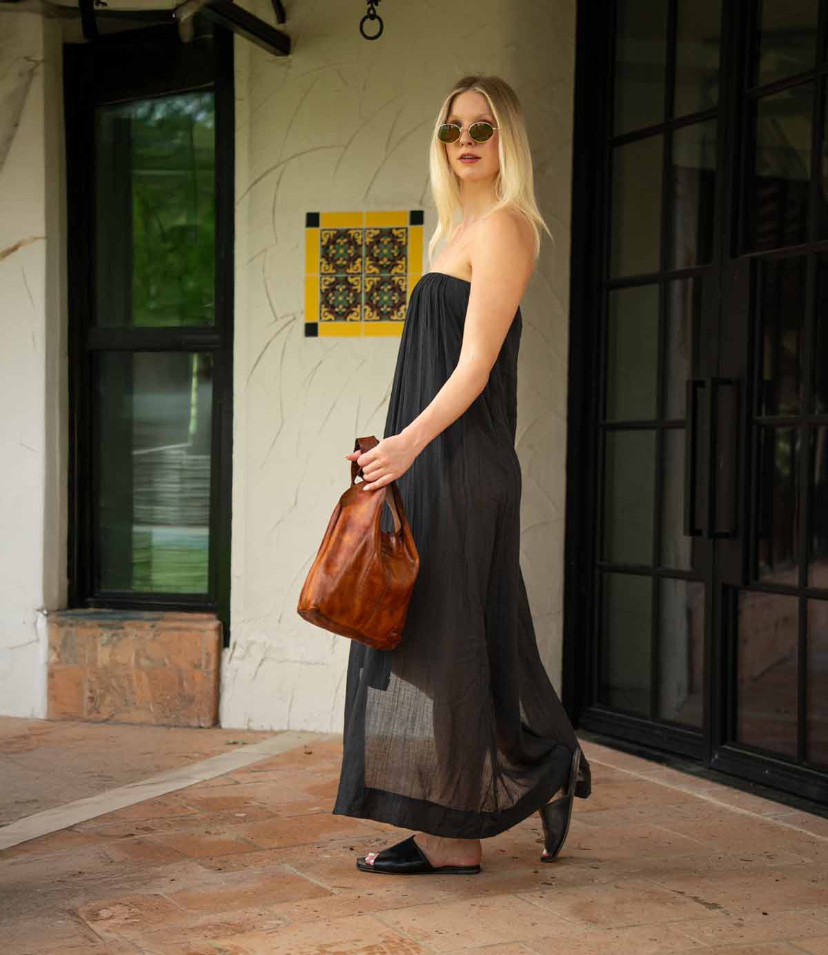 A woman in a black strapless dress, sunglasses, and sandals stands outdoors near glass doors, holding the Bed Stu Ariel Mid—an artisanal handbag crafted from vegetable-tanned leather.