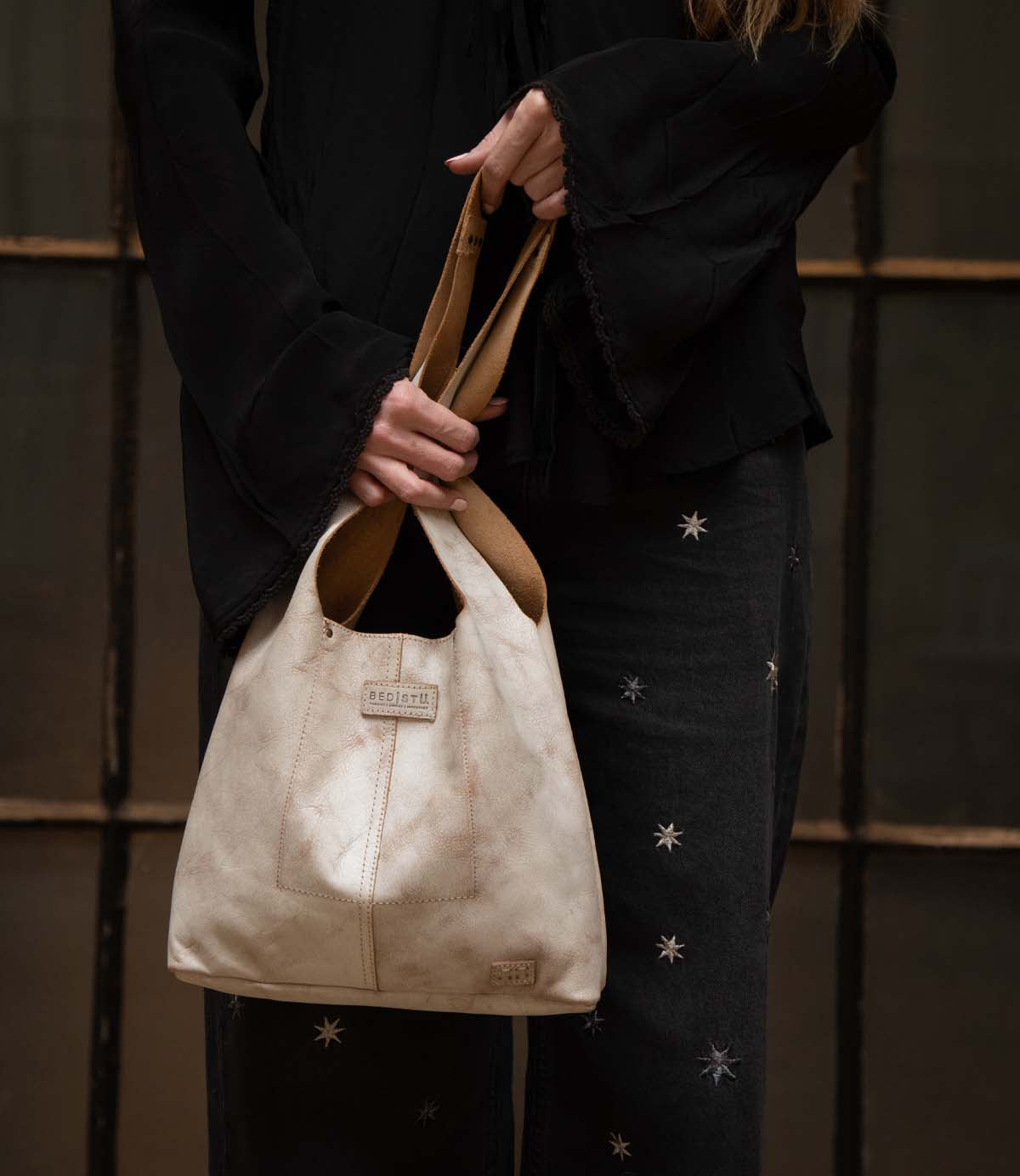 A person in a black top and star-patterned pants holds the Bed Stu Ariel Mid, a hand-finished leather bag with a magnetic closure, standing before a window with dark frames.