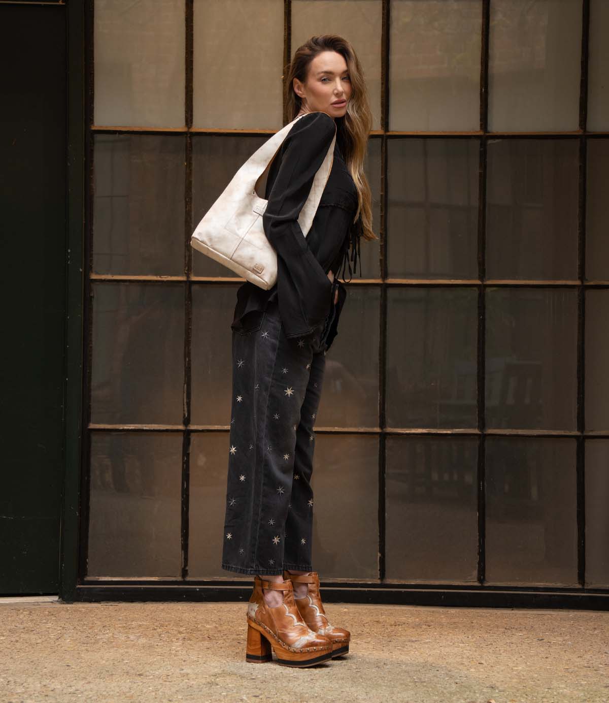 A woman stands sideways by large windows, wearing a black blouse, patterned black pants, brown platform shoes, and carrying the Bed Stu Ariel Mid—a large white hand-finished leather bag with a magnetic closure—over her shoulder.