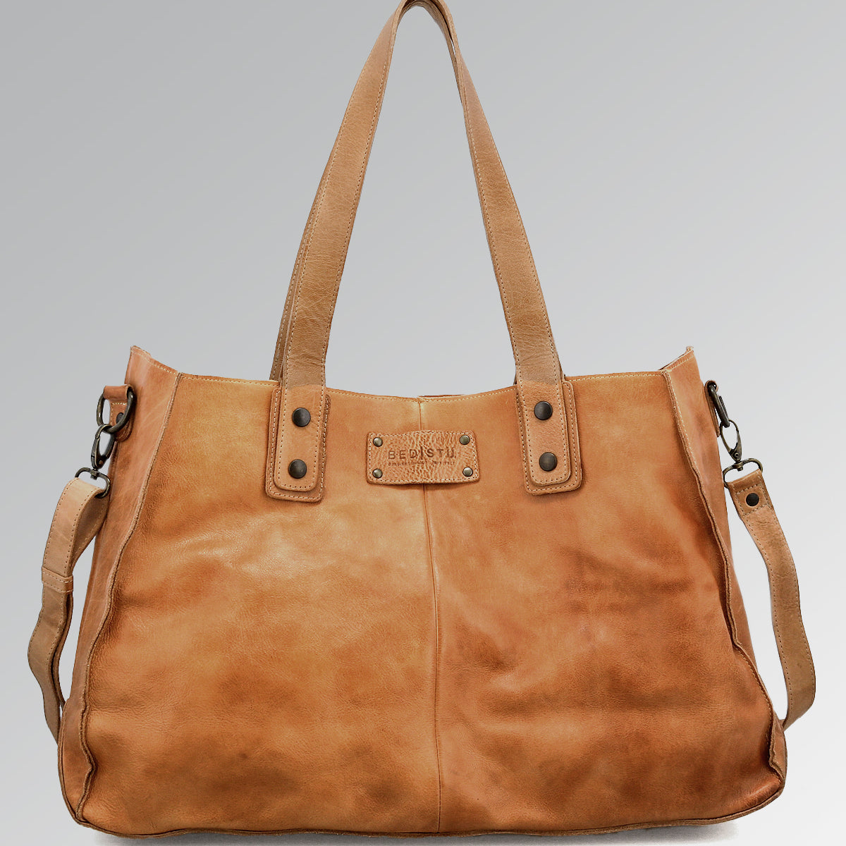 The Bed Stu Studio Arlington II Body is a large tan leather tote with two shoulder straps, a detachable crossbody strap, and a metal brand plate on the front, shown against a plain gray background.