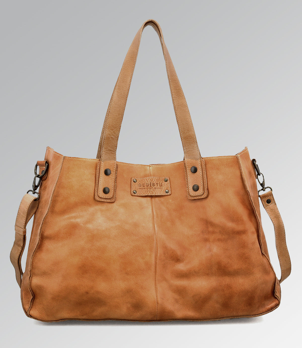 The Bed Stu Studio Arlington II Body is a large tan leather tote with two shoulder straps, a detachable crossbody strap, and a metal brand plate on the front, shown against a plain gray background.