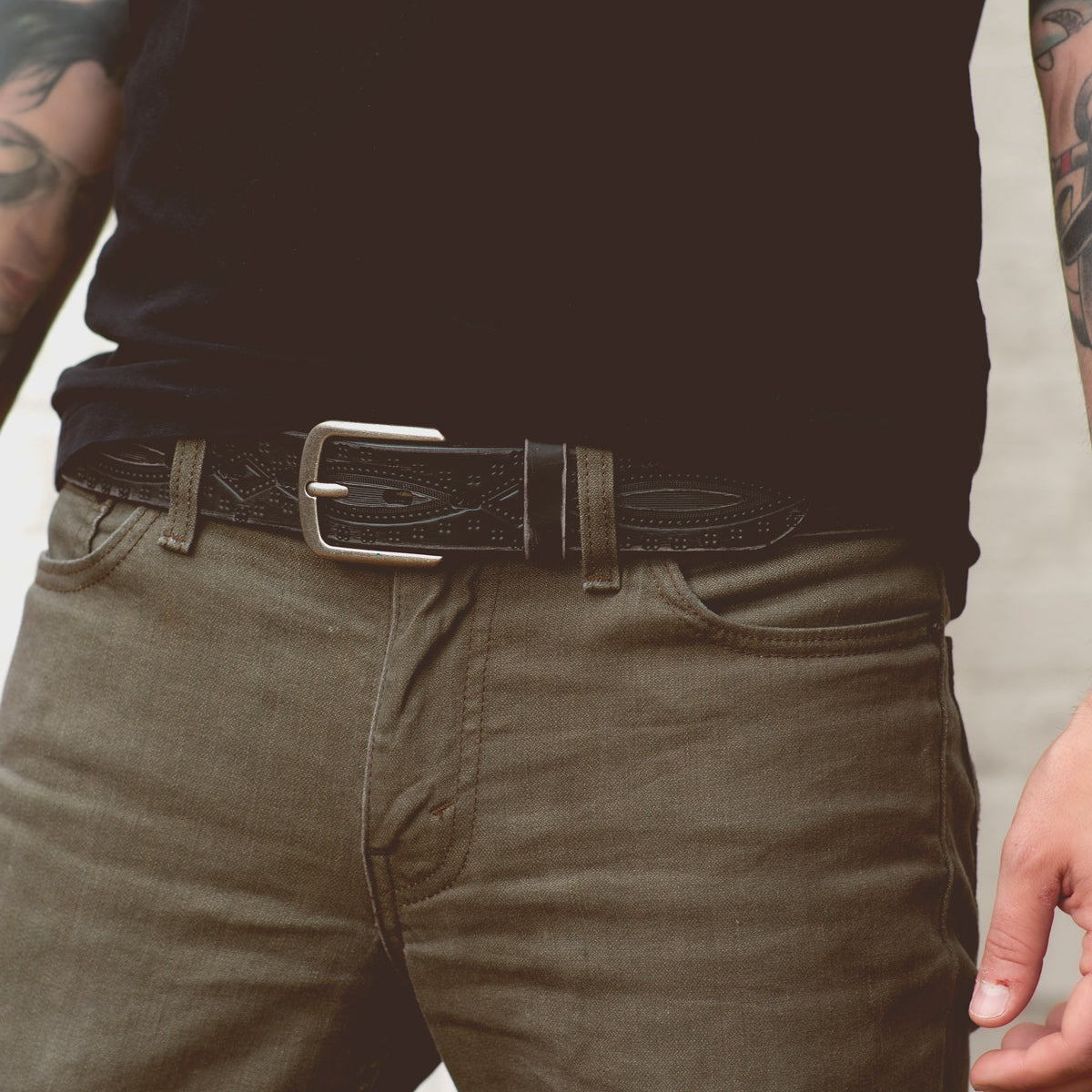 Man wearing a black shirt, dark jeans, and a Bed Stu Arsenal belt with a metal buckle adorned with western diamond details, and tattoos visible on his arms.