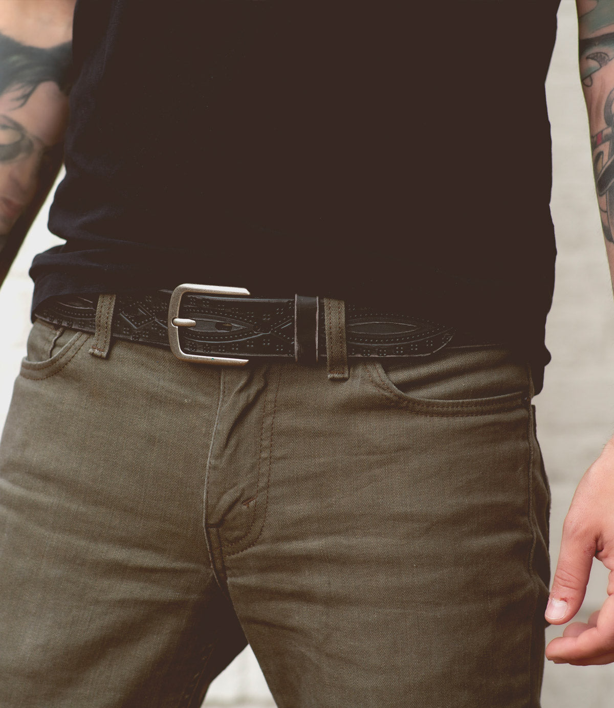Man wearing a black shirt, dark jeans, and a Bed Stu Arsenal belt with a metal buckle adorned with western diamond details, and tattoos visible on his arms.