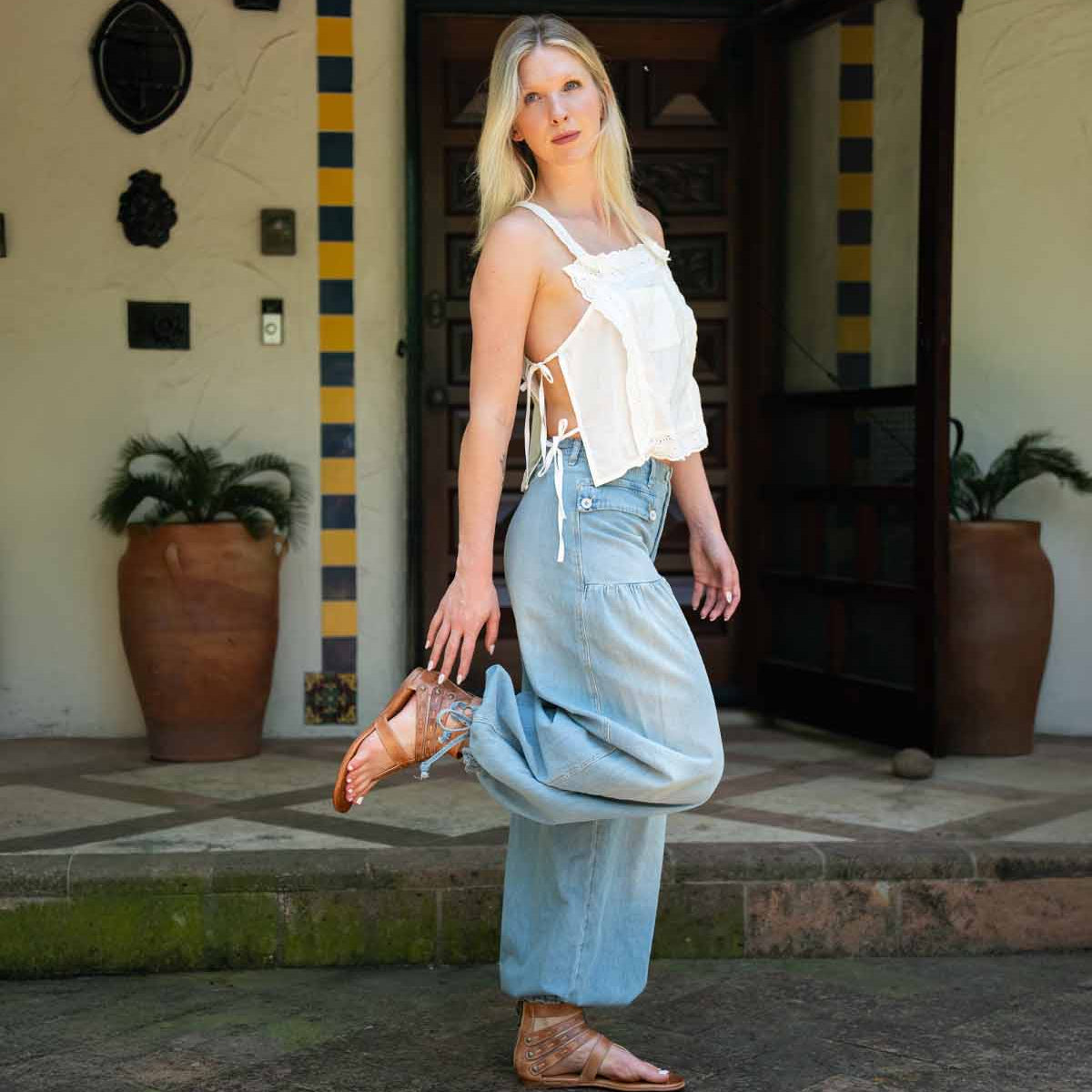 A woman stands outside a building in a white sleeveless top, light blue jeans, and Bed Stu’s Artemis II brown gladiator sandals with leather straps, one foot raised beside potted plants.