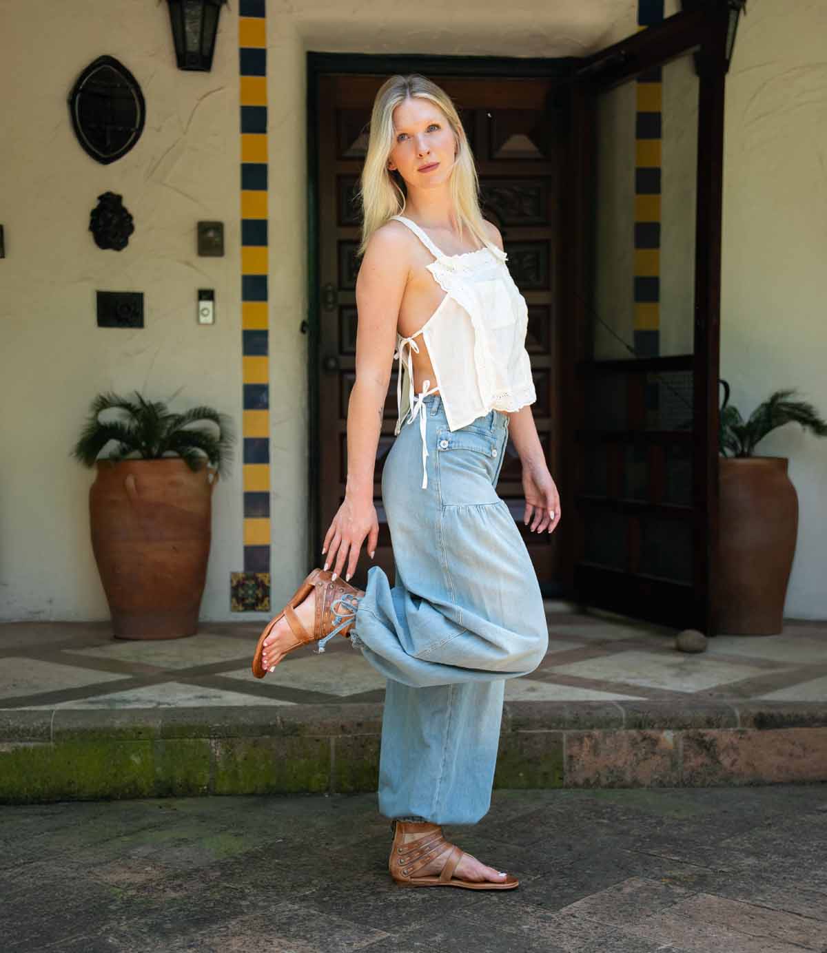A woman stands outside a building in a white sleeveless top, light blue jeans, and Bed Stu’s Artemis II brown gladiator sandals with leather straps, one foot raised beside potted plants.