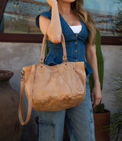 A woman in a denim vest and jeans stands by a cactus, carrying the Bed Stu Astana— a modern handbag inspired by vintage bowling bags with both shoulder and crossbody straps.