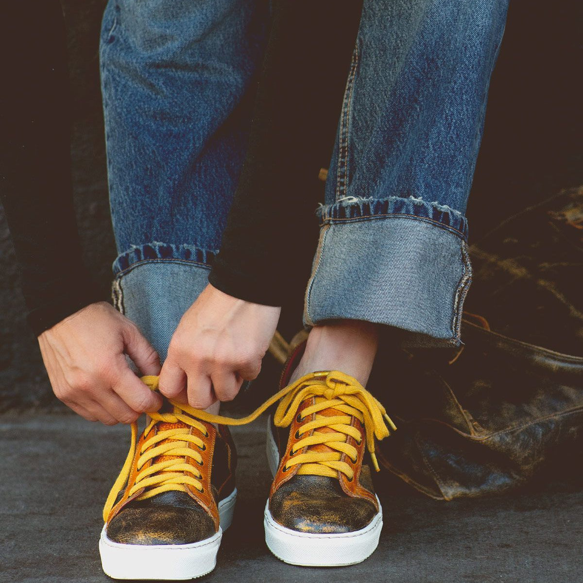 Person tying the yellow laces of Azeli sneakers by Bed Stu, featuring a sleek black and gold design, wearing rolled-up denim jeans and a black long-sleeve shirt—perfectly blending style with comfort.