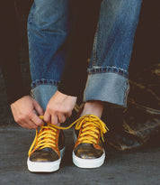 Person tying the yellow laces of Azeli sneakers by Bed Stu, featuring a sleek black and gold design, wearing rolled-up denim jeans and a black long-sleeve shirt—perfectly blending style with comfort.