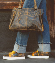 Person in cuffed jeans carrying a distressed Bed Stu Azeli leather handbag, wearing low-profile sneakers with yellow laces and a durable white outsole, where style meets comfort.