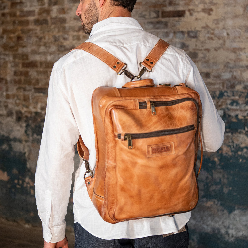 Person wearing a tan leather backpack with adjustable shoulder straps, front zipper pockets, and a top handle.