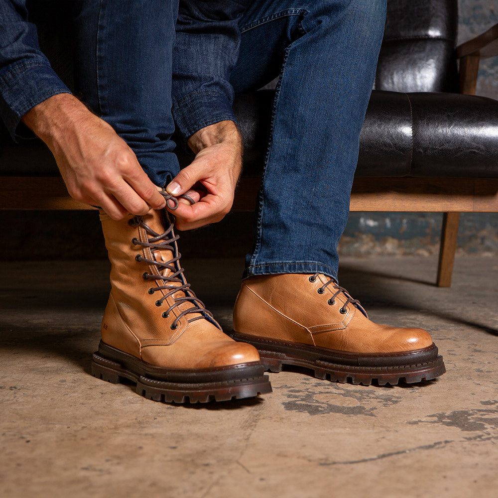 Person tying tan leather lace-up boots while wearing blue jeans.