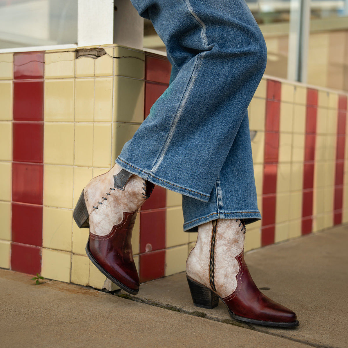 A person wearing blue jeans and Bed Stu Baila II Western-inspired boots with a pointed toe stands on a sidewalk next to a red and yellow tiled wall.