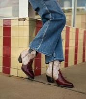 A person wearing blue jeans and Bed Stu Baila II Western-inspired boots with a pointed toe stands on a sidewalk next to a red and yellow tiled wall.