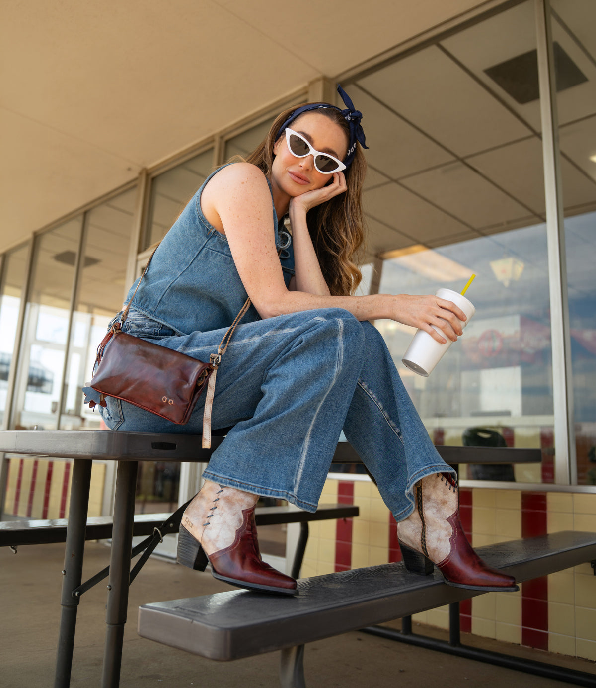A woman in a denim outfit and white sunglasses sits on a picnic table with a drink cup. She carries a brown purse, wears Bed Stu Baila II Western-inspired pointed-toe boots, and has a blue bandana in her hair.