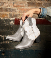 A hand holds the Bed|Stü Baila II boots—gray and white leather, Western-inspired with intricate stitching—set against a worn brick and painted wall background.