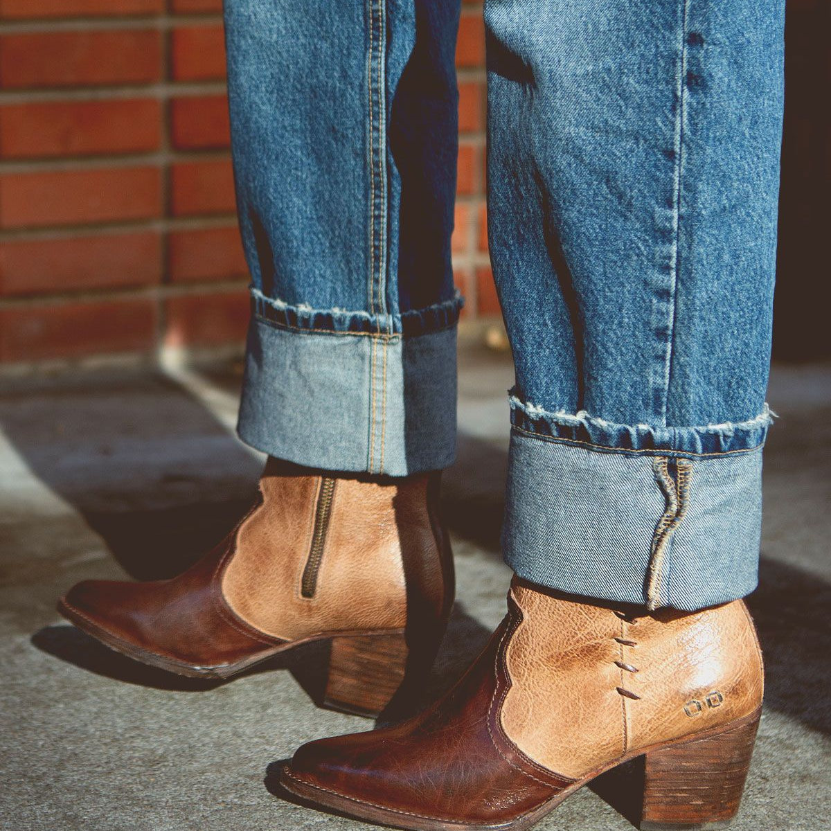 A person wearing rolled-up blue jeans and the Bed Stu Baila II brown leather ankle boots with chunky heels and whipstitch accents stands on a concrete surface against a brick wall backdrop, exuding cowgirl vibes.