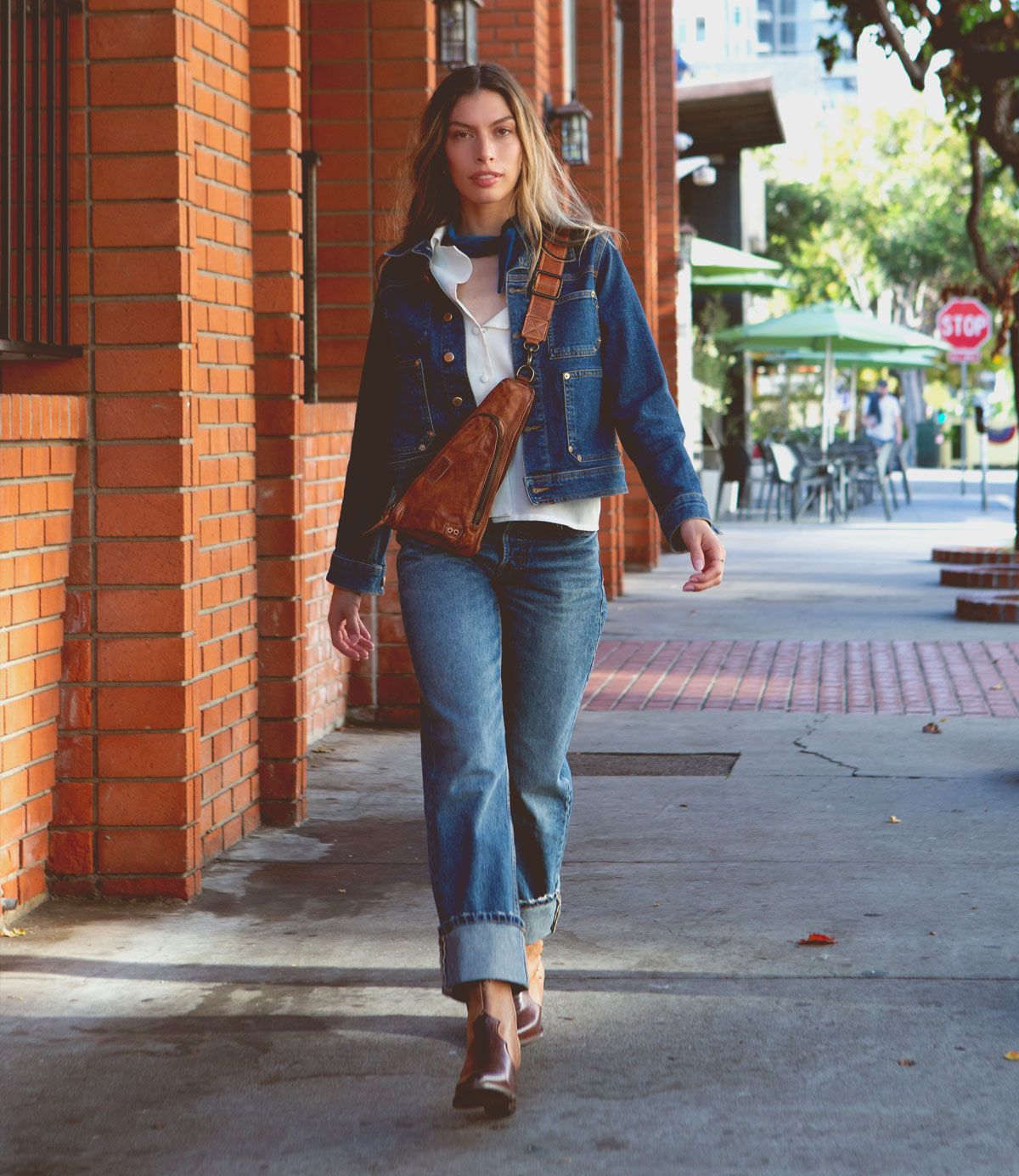 Person walking down a sidewalk in a denim outfit, carrying the Bed Stu Baila II crossbody bag, exuding cowgirl vibes. Brick building and outdoor seating area visible in the background.