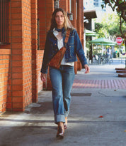 Person walking down a sidewalk in a denim outfit, carrying the Bed Stu Baila II crossbody bag, exuding cowgirl vibes. Brick building and outdoor seating area visible in the background.