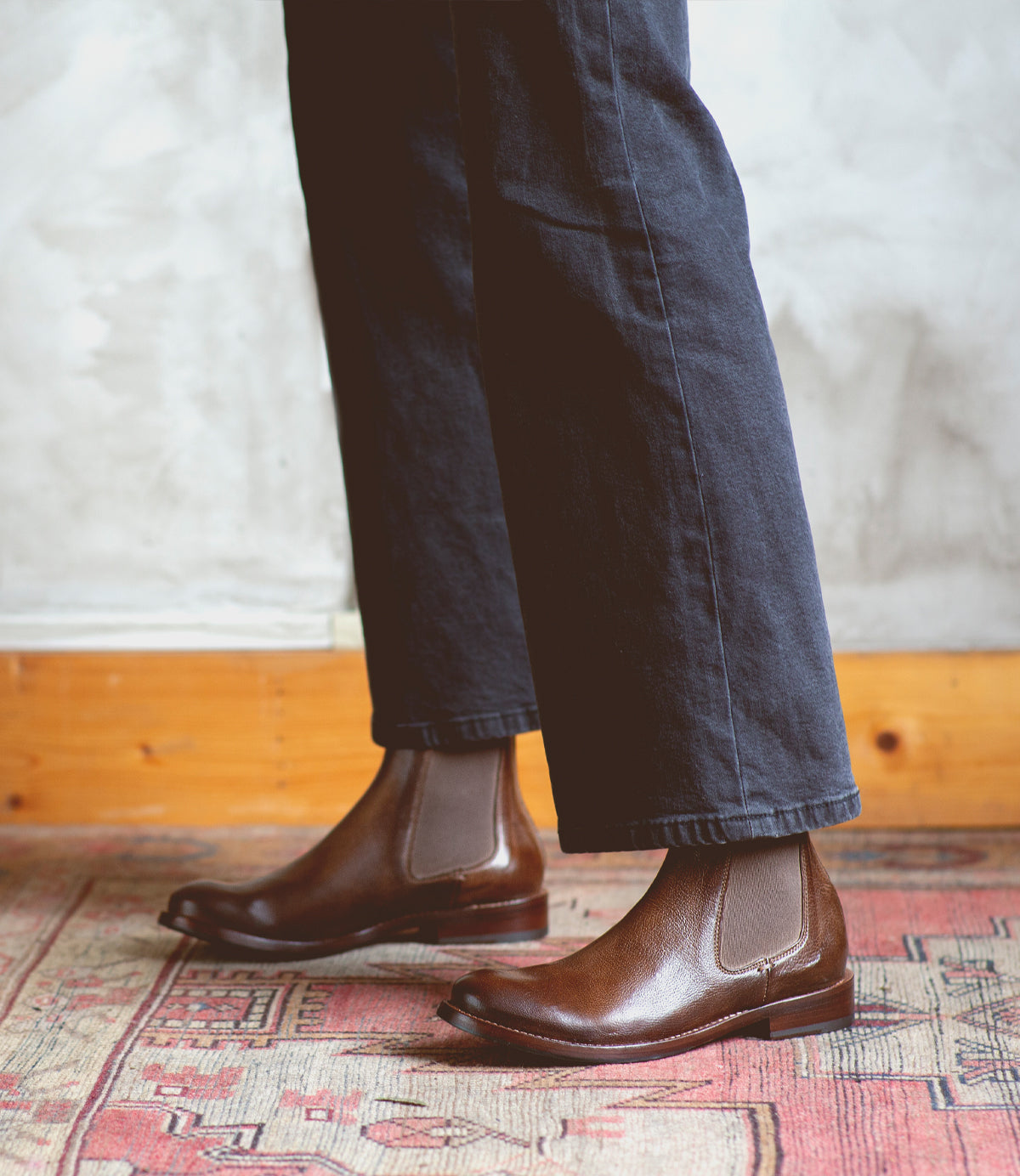 A person standing on a Barfly rug wearing brown Bed Stu Chelsea boots made of Italian leather.