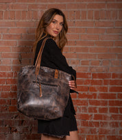 A woman in a black dress stands by a brick wall, carrying the Bed Stu Barra II, a large handcrafted tote bag with an organic cotton lining, over her shoulder.