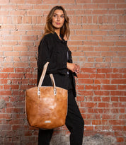 A person in black stands against a brick wall, carrying the Bed Stu Barra II—a large handcrafted tote bag with an organic cotton lining—over their shoulder.