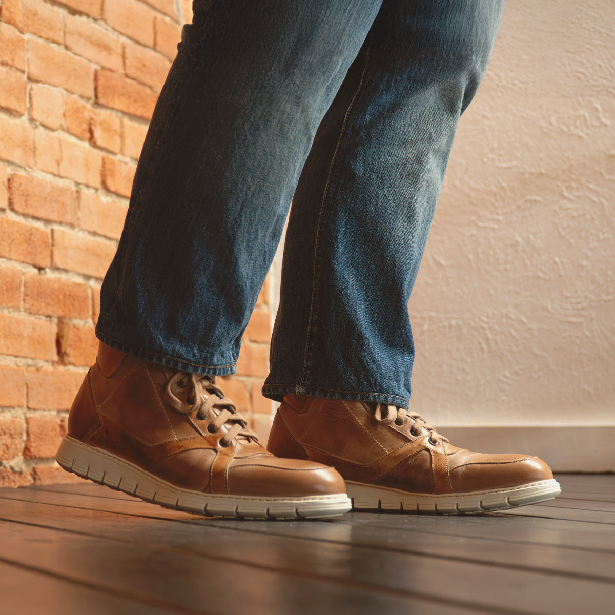 A person stands on a wooden floor next to a brick wall, wearing Bed Stu's Barton brown leather high-top sneakers with blue jeans.
