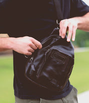 A person in a black shirt is unzipping the durable strap of a Beau black leather sling backpack by Bed Stu outdoors.