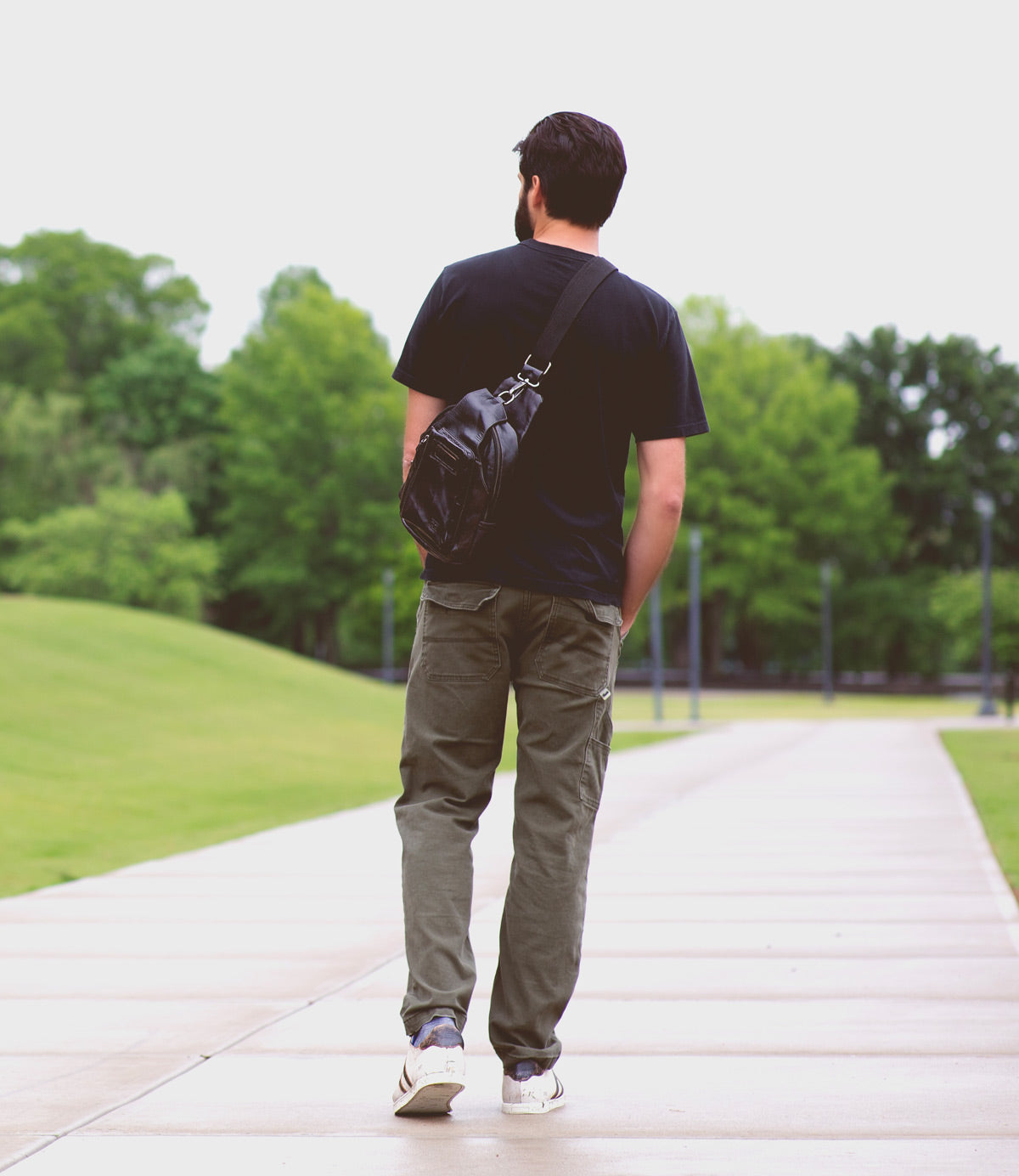 A man with a beard walks along a paved path in a park, wearing a black shirt, khaki pants, white sneakers, and carrying the Bed Stu Beau leather sling backpack with a durable strap. Green trees and grass line the background.