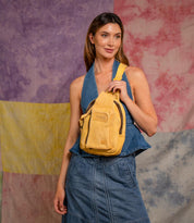A woman in a blue denim halter top and skirt poses with the Bed Stu Beau compact hands-free backpack in front of a tie-dye background.