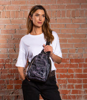A woman stands against a brick wall, wearing a white t-shirt and black pants, carrying the Bed Stu Beau sleek leather sling backpack for a stylish hands-free option.