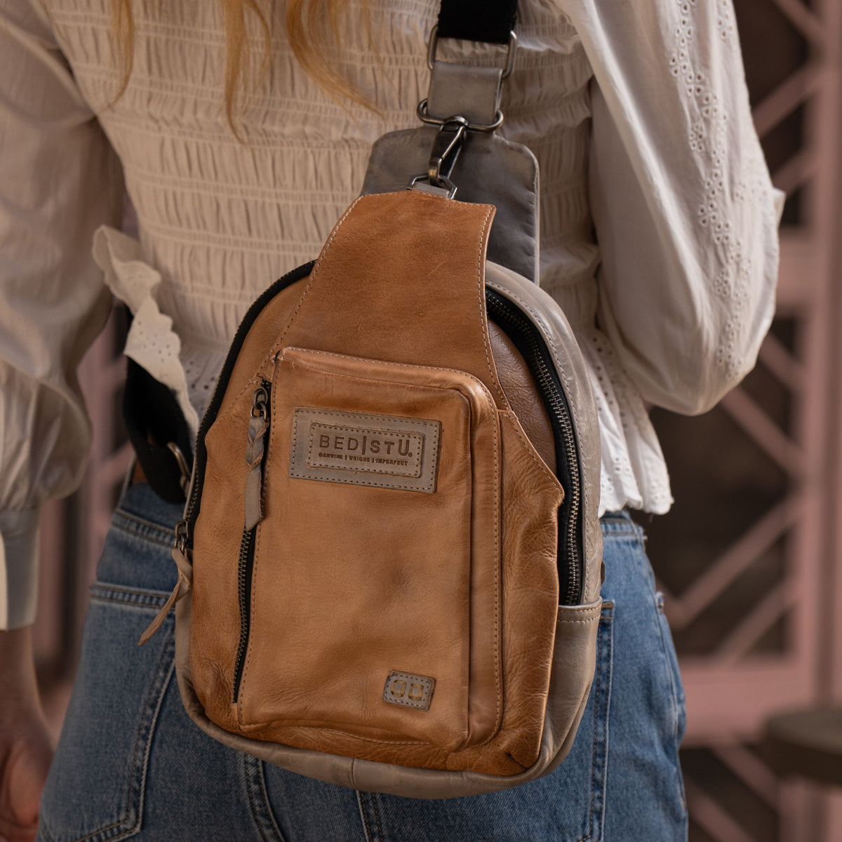 Wearing the Bed Stu Beau tan leather crossbody bag, a stylish unisex sling with a front zipper pocket, paired with a white blouse and blue jeans.