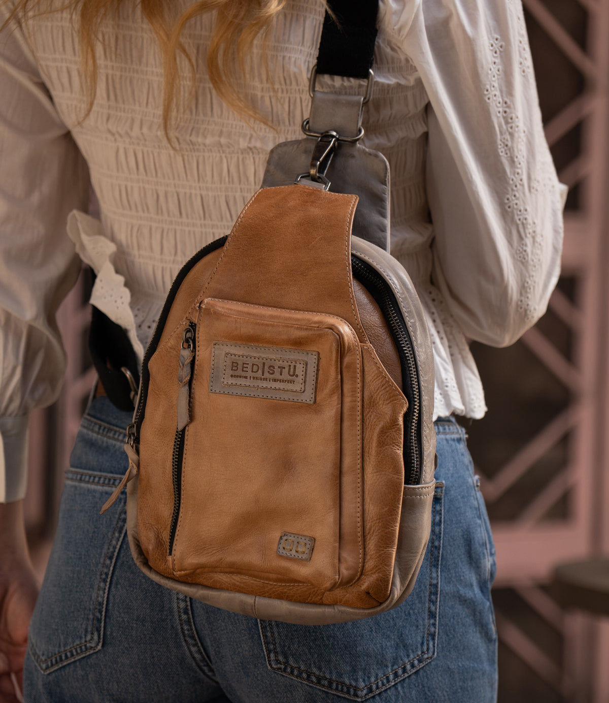 Wearing the Bed Stu Beau tan leather crossbody bag, a stylish unisex sling with a front zipper pocket, paired with a white blouse and blue jeans.