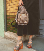 A person standing against a yellow-brick wall, holding a Bed Stu Beau brown leather sling backpack with an outer zipper pocket, wearing a black skirt and brown heeled sandals.