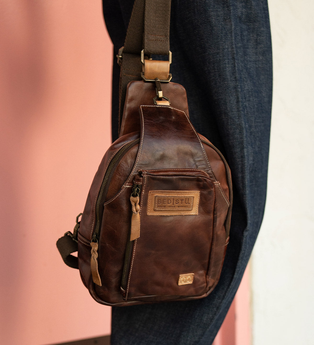 The Beau sling backpack by Bed|Stü, crafted in brown leather with a zipper pocket and a tan patch label, hangs on a person's shoulder against a pink and white background.