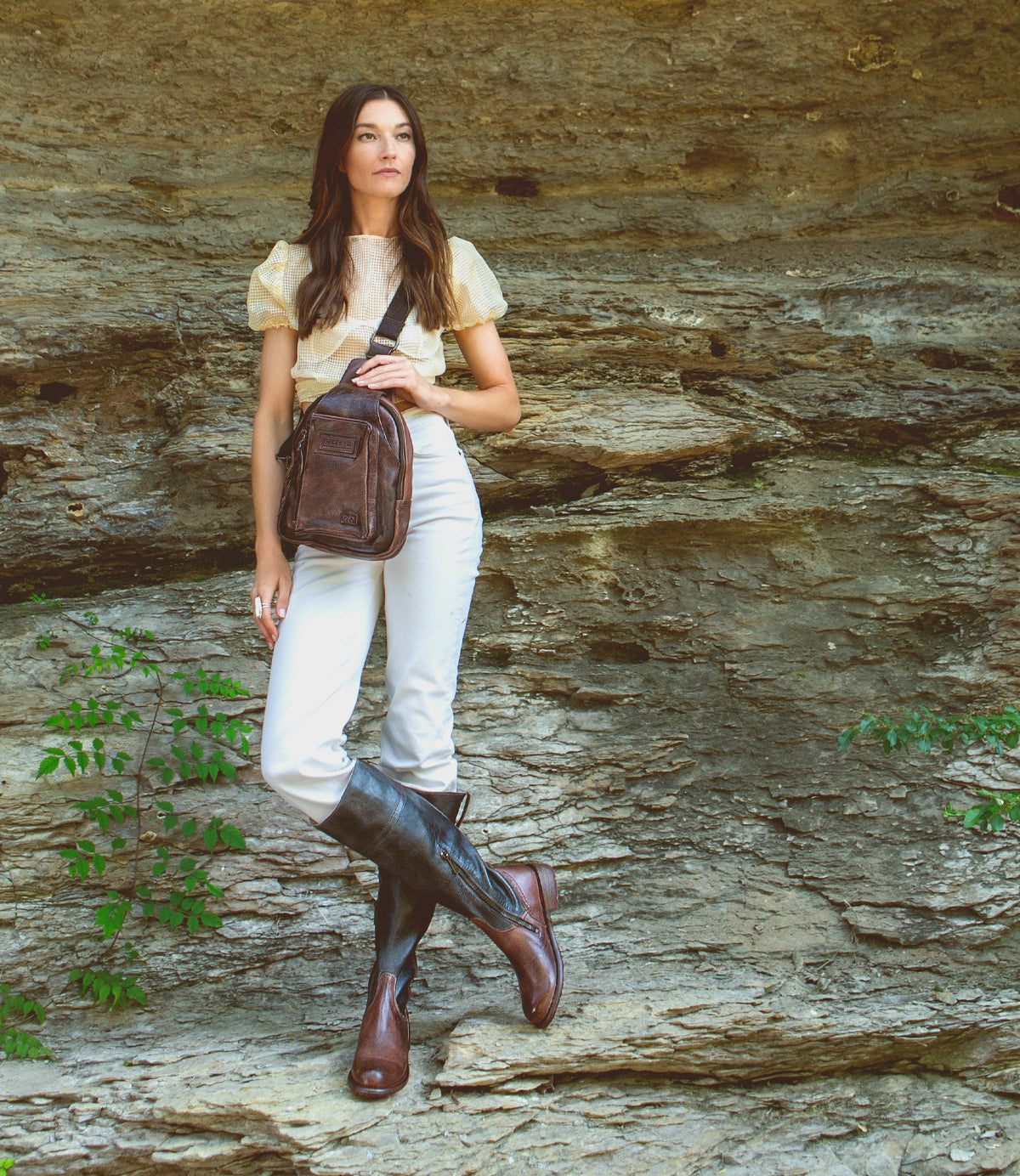 A woman is standing on a rock with a Beau sling backpack from Bed Stu, which features a durable strap holding the leather backpack.