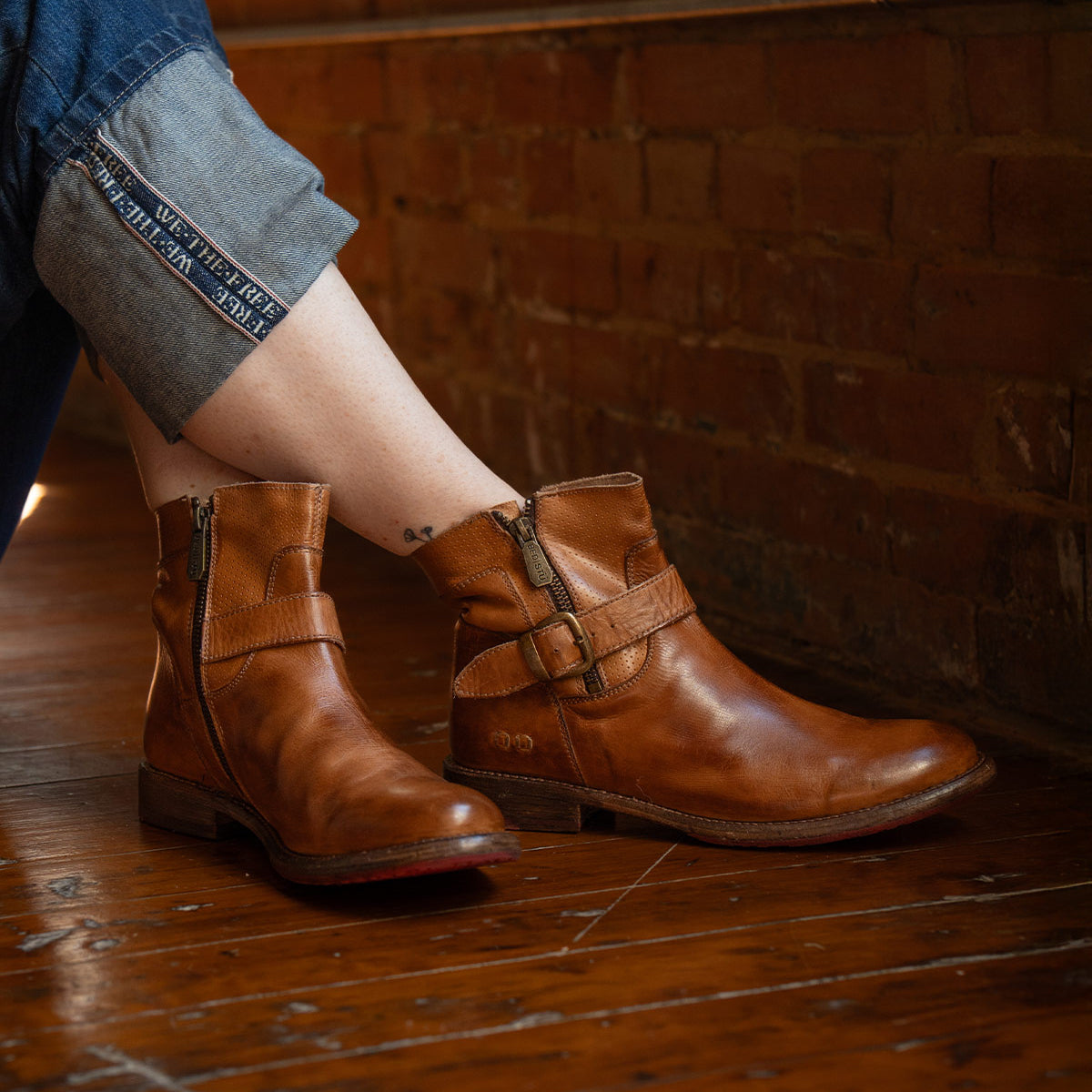A person in rolled-up jeans and Bed Stu's Becca brown leather short engineer boots with dual zippers sits on a wooden floor against a brick wall.