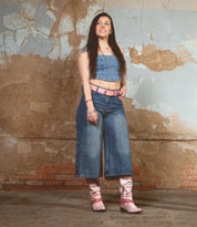 A woman in a denim crop top and jeans stands against a textured wall, wearing pink Bed Stu Western boots named "Betting," featuring decorative harness straps, along with a matching pink belt.