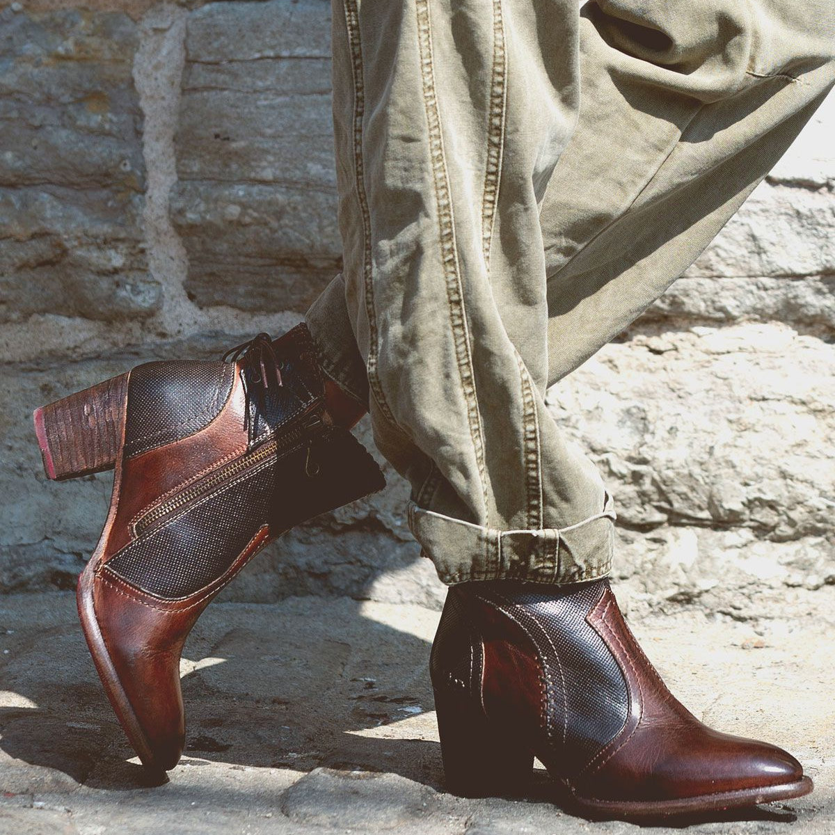 A person wearing Bia brown leather booties made from vegetable-tanned leather by Bed Stu and khaki pants stands near a stone wall, embodying all-day wear comfort.
