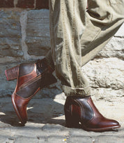 A person wearing Bia brown leather booties made from vegetable-tanned leather by Bed Stu and khaki pants stands near a stone wall, embodying all-day wear comfort.