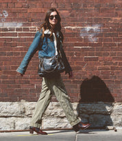 A person wearing sunglasses, a denim jacket, green pants, and brown leather booties walks by a brick wall carrying the Bia bag by Bed Stu.