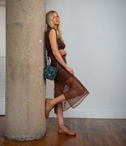 A woman in a sheer brown dress and brown shoes—the Bingo II by Bed|Stü, featuring an EXTRALIGHT outsole—leans against a large concrete pillar indoors, holding a dark satchel bag and standing on a wooden floor.