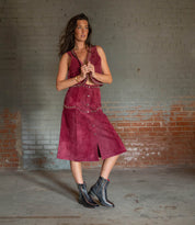 A woman stands indoors by a faded brick wall, wearing the Bed Stu Bitsy sleeveless maroon dress with metallic studs and black ankle boots with a stacked wooden heel.